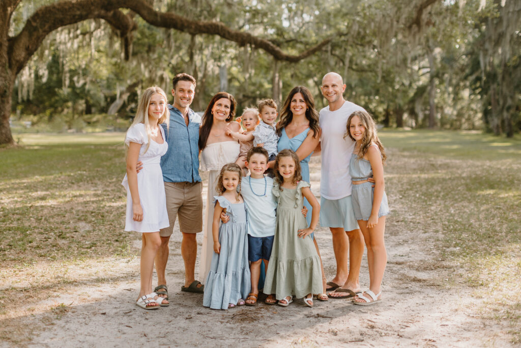 family posing outdoors under a large oak tree with Spanish moss, dressed in coordinated pastel and neutral tones, smiling in natural sunlight.