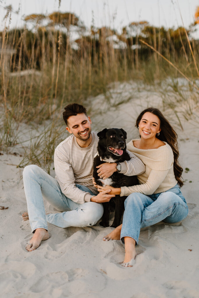 A couple with light skin and a black dog sit together on a sandy Hilton Head beach at sunset, surrounded by tall grass. They are dressed in casual, neutral-toned clothing, with the man wearing a striped sweater and light jeans, and the woman wearing an off-the-shoulder cream sweater and blue jeans. They are both barefoot, smiling warmly, and looking happy. The dog, sitting between them, has a content expression with its tongue out. The golden light and soft focus on the background create a warm, intimate atmosphere.