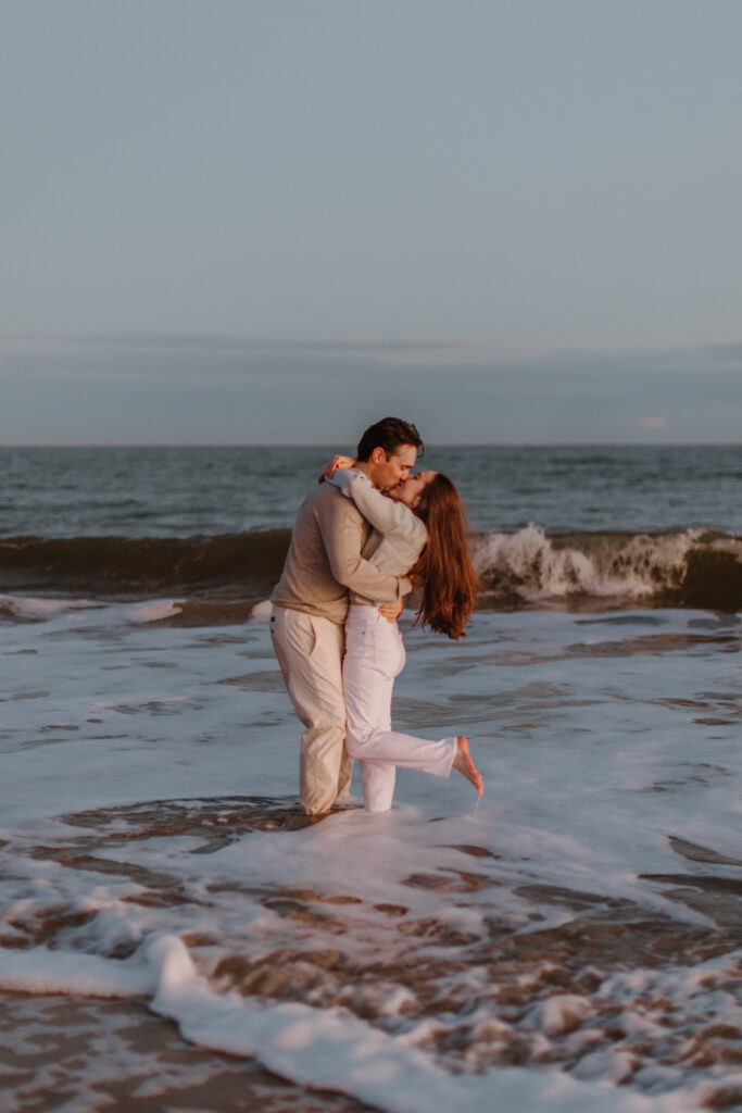 A couple shares a romantic kiss while standing in the ocean waves during a Hilton Head sunset. The man, dressed in a light sweater and pants, holds the woman close as she wraps her arms around his neck. She wears a cozy sweater and white pants, lifting one leg playfully as they embrace. The golden light of the setting sun casts a warm glow on the scene, with gentle waves rolling in the background.