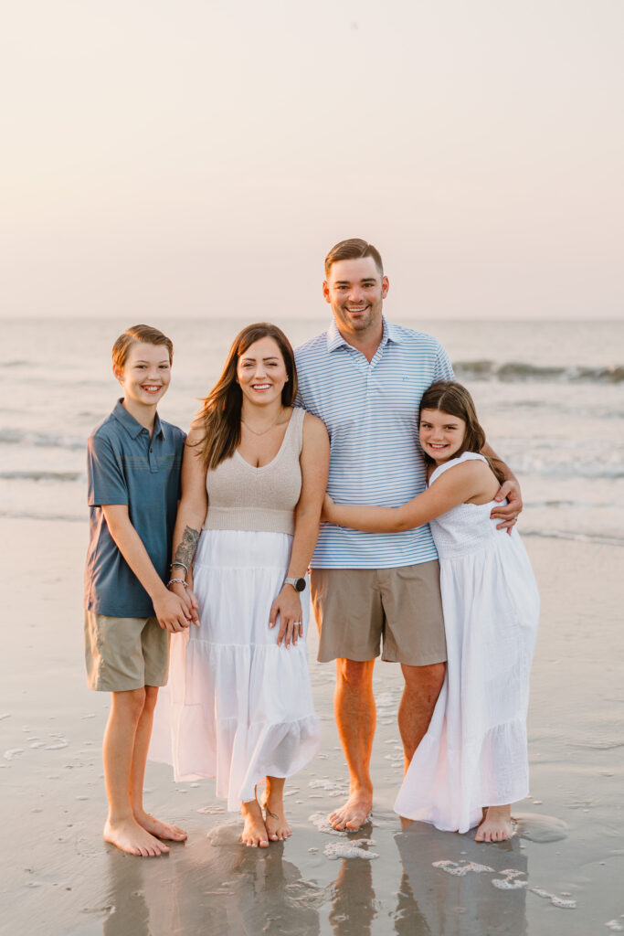 A family of four stands barefoot on a Hilton Head beach at sunrise, smiling at the camera. The mother, wearing a white flowy dress and a beige knit top, holds hands with her son, who is dressed in a navy polo and khaki shorts. The father, in a striped polo and tan shorts, stands beside them with his daughter hugging his waist. The daughter wears a white dress, mirroring her mother’s style. The ocean waves and soft golden light create a warm and serene backdrop.