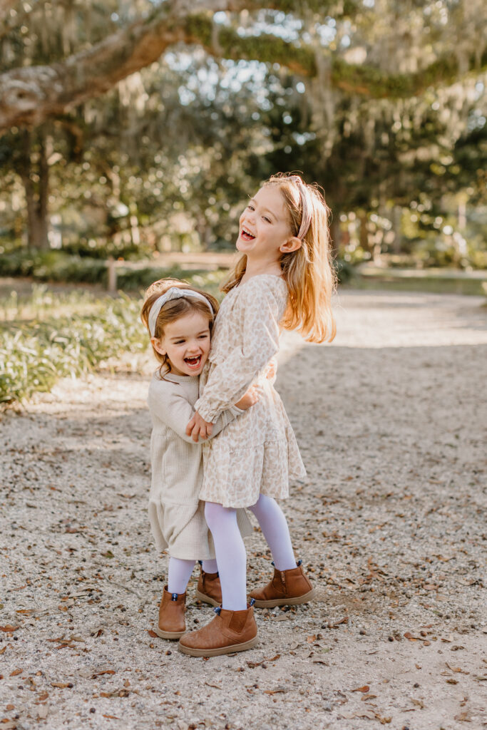 Hilton Head family photographer captures two young sisters laughing and hugging in a sunlit outdoor setting with moss-draped trees in the background.