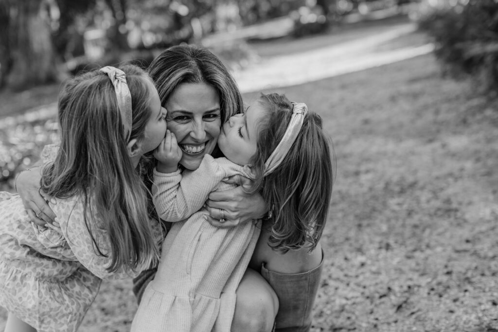 Hilton Head family photographer captures a joyful mother embracing her two daughters as they kiss her cheeks in a candid outdoor portrait.